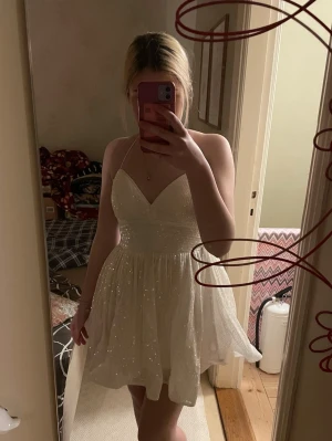 Glittrig vit kortklänning - Supersöt vit kortklänning med glitter och v-ringning. Klänningen har tunna axelband som korsas i ryggen och snörning baktill för en snygg passform. Perfekt till t.ex. student!