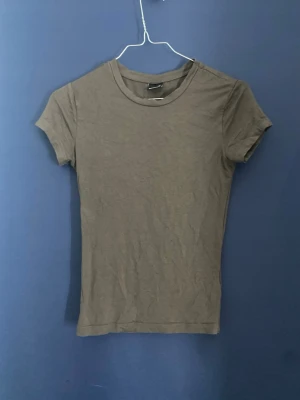 Mörkgrå basic t-shirt från H&M - En enkel mörkgrå t-shirt från gina tricot med rund halsringning och korta ärmar. Den är väldigt stretchig som känns skön mot huden. Perfekt att matcha med jeans eller shorts för en clean och avslappnad stil.