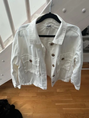 Vit jeansjacka med bruna knappar - Snygg vit jeansjacka från Bubbleroom med klassisk krage, två stora bröstfickor och bruna knappar framtill. Jackan har en rak passform och är tillverkad i denim. Perfekt att slänga över en hoodie eller t-shirt för en cool look.