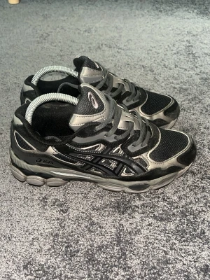 Svarta och grå Asics sneakers - Snygga sneakers från Asics i svart och grått med coola detaljer och meshpaneler för extra andningsförmåga. Skorna har en chunky sula och klassiska Asics-loggan på sidan. Perfekta för dig som gillar sportig och trendig stil.