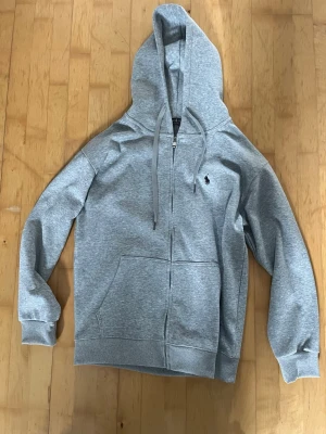 Polo Ralph Lauren  - Snygg grå hoodie med dragkedja från Polo Ralph Lauren. Klassisk design med huva och dragsnören, två stora fickor fram och den ikoniska svarta polospelaren broderad på bröstet. Mjuk bomullsblandning som är skön att ha på sig. Knappt använd. Skriv för frågor eller fler bilder. Passar både M och L