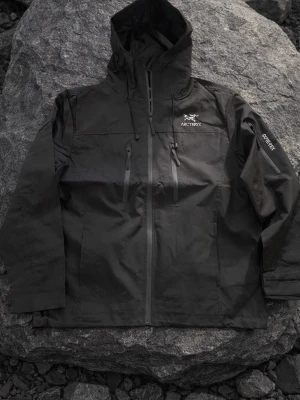 Svart vindjacka från Arc'teryx - Snygg svart vindjacka från Arc'teryx med dragkedja framtill, två bröstfickor med vattentäta dragkedjor och justerbar huva. Jackan är tillverkad i slitstarkt material och har Gore-Tex-tyg för extra väderskydd. Perfekt för dig som gillar outdoor och streetwear. Använd ett par gånger inga märken storlek M