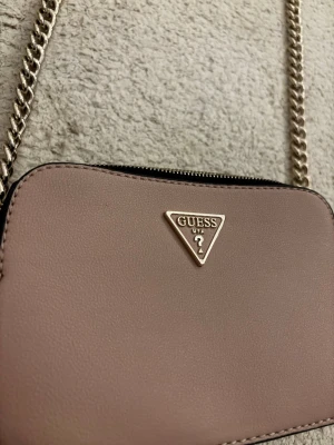 Rosa axelväska  (inte äkta Guess) - Snygg rosa axelväska från Guess med guldfärgad kedjerem och klassisk trekantslogga framtill. Väskan har en stilren, rektangulär form och är tillverkad i slätt konstläder. Perfekt för dig som vill ha en trendig och enkel accessoar.