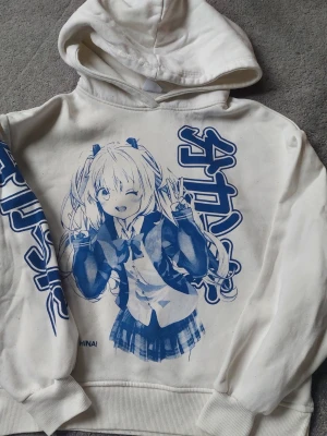 Vit hoodie med blå anime-tryck - Säljer en vit hoodie med stor blå anime-illustration framtill och japansk text på ärmen. Hoodien har huva och ribbade muddar. Perfekt för dig som gillar manga eller japansk popkultur. En del noppor som går att tabort.