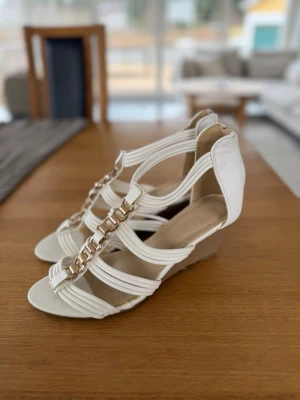 Vita sandaletter med guldkedja - Vita sandaletter med remmar och guldfärgad kedja framtill. Öppen tå och låg kilklack.