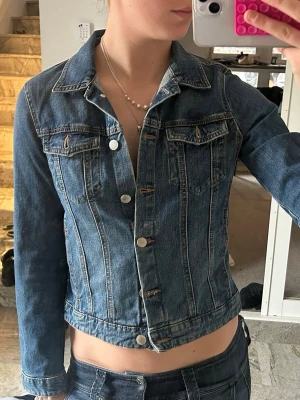 Mörkblå klassisk jeansjacka - Snygg mörkblå jeansjacka med figurnära passform och silverfärgade knappar. Jackan har två bröstfickor med lock och knapp samt markerade sömmar framtill och baktill. Perfekt att slänga över en t-shirt för en cool look.