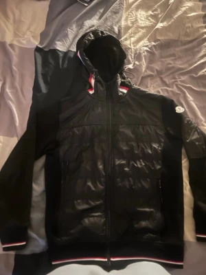 Svart Moncler vindjacka med huva - Snygg svart vindjacka från Moncler med huva och dragkedja framtill. Jackan har quiltade partier på framsidan, ribbade muddar med rödvita detaljer och Moncler-logga på ärmen. Perfekt för dig som vill ha en stilren och sportig look.