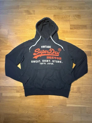 Superdry hoodie - Säljer en riktigt snygg svart superdry hoodie med röd och vit tryck, den mest efterfrågade färgkombinationen. Inga defekter och passar vanlig L. Hör av dig vid frågor eller prisförslag!