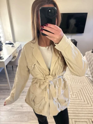 Beige kavaj med knytskärp - Stilren beige kavaj med knytskärp i midjan och två bröstfickor med lock. Kavajen har klassisk krage och är tillverkad i ett mjukt tyg som ger en avslappnad men trendig look. Perfekt att slänga över en t-shirt för en snygg outfit.