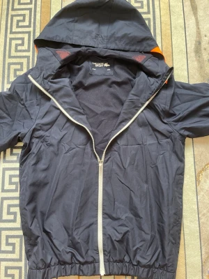 Svart vindjacka från Lacoste jätte fin  - Snygg svart vindjacka från Lacoste Sport med huva och dragkedja. Jackan har en orange detalj på axeln och klassisk Lacoste-logga på bröstet. Tillverkad i lätt och smidigt material, perfekt för blåsiga dagar. Varan är i jätte fin skick.  Frågor och funderingar kan ni gärna höra av er kom gärna med prisförslag också. 