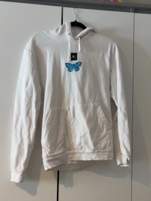 Vit hoodie med blå fjärilstryck - Snygg vit hoodie med stor blå fjäril på ryggen och en mindre fjäril på bröstet. Hoodien har huva med dragsko och en klassisk känguruficka framtill. Perfekt för dig som gillar unika tryck och vill sticka ut lite extra. Mjuk bomullskänsla. Säljs då den inte kom till användning.