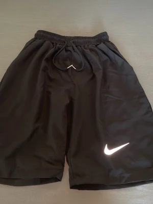 Svarta sportshorts från Nike - Säljer ett par svarta sportshorts från Nike med elastisk midja och dragsko. Shortsen har en klassisk passform och är tillverkade i ett lätt och snabbtorkande material. Nike-loggan syns tydligt på ena benet. Perfekta för träning eller chill.