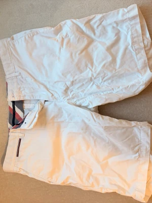 Vita shorts från Tommy hilfiger - Säljer ett par klassiska vita shorts från Tommy hilfiger med diskret rutigt innerfoder i midjan och knappstängning. Perfekta för sommardagar och enkel att matcha.