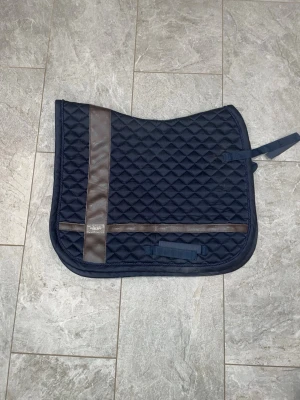 Marinblått schabrak med bruna detaljer - Säljer ett marinblått schabrak med rutmönster och bruna detaljer. Schabraket har en quiltad yta och en dekorativ brun rand. Perfekt till hästutrustning och ridning.