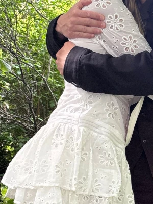 Vit broderad kortklänning med volang - Söt vit kortklänning med broderade blommor och volangdetaljer. Klänningen har markerad midja och är tillverkad i ett lätt tyg som passar perfekt för varma dagar. Ärmarna är långa och klänningen har en romantisk känsla.