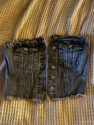 Mörkblå denim tubtopp med knappar - Trendig tubtopp i mörkblå denim med fransig kant, dekorativa knappar framtill och två bröstfickor. Baksidan har smock för skön passform. Perfekt för dig som vill ha en edgy och unik look.
