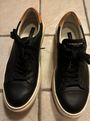 Sneaky Steve svarta lädersneakers - Svarta sneakers i läder från Sneaky Steve med vit tjock sula och bruna detaljer vid hälen. Klassisk låg modell med svarta skosnören och stilren design. Perfekta för dig som gillar en clean och modern look.