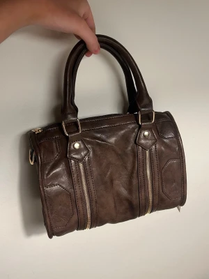 Brun handväska med dubbla dragkedjor - Snygg brun handväska i läderimitation med två guldfärgade dragkedjor framtill och matchande gulddetaljer