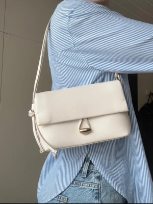 Benvit handväska med guld detaljer från mango - Benvit handväska från Mango med magnetstängning och dekorativt spänne framtill. Axelbandet går att justera med hjälp av en knyt på ena sidan. Guldiga detaljer och två fack inuti. Ensams använd en gång.  Mått: 23x15x8 cm.  Nypris 529 kr.