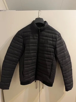 Svart pufferjacka från Armani Exchange - Snygg svart pufferjacka från Armani Exchange med diskret logotyp på bröstet. Jackan har ståkrage, dragkedja framtill och quiltad design. Perfekt för kalla vinterdagar och ger en stilren look.