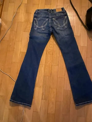 Blå bootcut jeans med broderade fickor - Snygga blå bootcut jeans med vita kontrastsömmar och broderade detaljer på bakfickorna. Jeansen har låg midja och klassisk femficksdesign. Perfekta för dig som gillar en retroinspirerad look och vill sticka ut lite extra. Har använt dom fåtal gånger💕