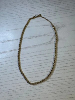 Guldfärgat tvinnat halsband - Snyggt guldfärgat halsband med tvinnad kedja. Halsbandet har en enkel och stilren design med karbinlås och liten rund bricka vid låset. Perfekt för att ge din outfit en elegant touch.