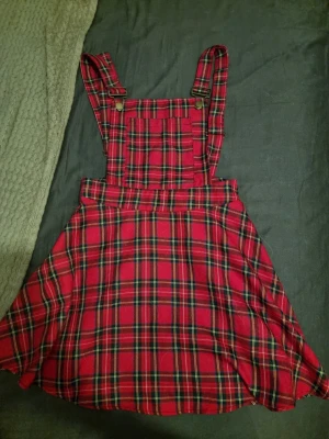 Röd rutig hängselkjol från Jawbreaker - Supersnygg röd hängselkjol från Jawbreaker i klassiskt rutigt mönster. Kjolen har justerbara axelband med metallspännen och en ficka framtill på bröstet. Perfekt för dig som gillar alternativ stil och vill sticka ut lite extra.