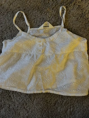 Vit broderad linnetopp från H&M - Söt vit linnetopp från H&M med broderade blommor och tunna axelband. Toppen har dekorativa knappar framtill och en scallop-kantad urringning. Perfekt för varma dagar och enkel att styla med jeans eller kjol. Är i barn stolek!