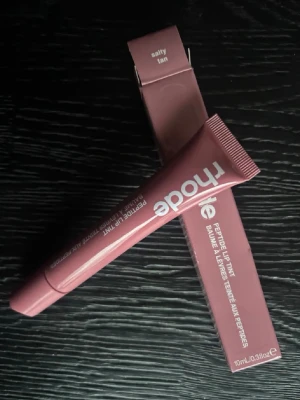 Rhode Peptide Lip Tint Salty Tan - Rhode Peptide Lip Tint i färgen Salty Tan ger läpparna en mjuk brunrosa ton. Läppglanset kommer i en smidig tub med vit text och rymmer 10 ml. Formulan är berikad med peptider för extra vård och glans. Perfekt för dig som vill ha en naturlig look med lite extra glow.