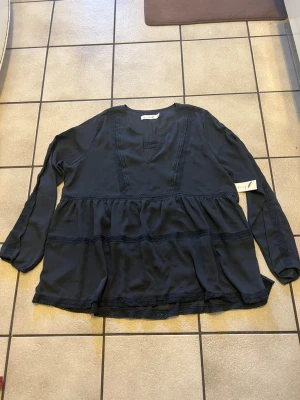 Svart blus med spetsdetaljer storlek M - Snygg svart blus med v-ringning och dekorativa spetsdetaljer längs framstycket och nederdelen. Blusen har en lös passform och långa ärmar, perfekt för en trendig och avslappnad look. Tillverkad i ett mjukt och lätt tyg.