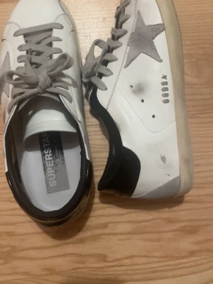 Golden Goose Superstar vita sneakers - Golden Goose Superstar sneakers i vitt läder med grå mockastjärna på sidan, grå skosnören och svart häl. Klassisk låg modell med vintage-inspirerade detaljer och texten 'SUPERSTAR' på innersulan. Perfekta för en cool och avslappnad stil.
