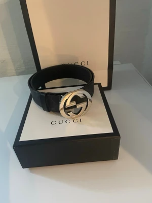 Svart Gucci bälte  - Snyggt svart bälte från Gucci med klassiskt GG-spänne i silver med original förpackning och kvitto. Bältet har diskret mönster längs remmen och är tillverkat i canvas. Perfekt accessoar för att lyfta din outfit med en lyxig touch.                                                                  OBS pris kan diskuteras vid snabb affär.