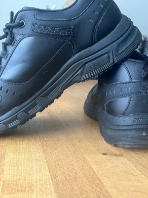 Svarta Skechers sneakers i läder - Stilrena svarta sneakers från Skechers i läder med robust sula och klassisk snörning. Skon har diskret logga på sidan och detaljerade sömmar för en sportig look. Perfekta för dig som gillar en clean och enkel stil.