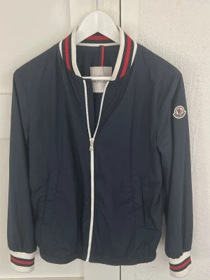 Marinblå vindjacka från Moncler - Snygg marinblå vindjacka från Moncler med vit dragkedja och ribbade muddar i vitt, rött och marinblått. Jackan har två sidofickor och klassisk Moncler-logga på ärmen. Tillverkad i ett lätt, glansigt material som ger en sportig och fräsch look. Jackan är sparsamt använt och i fint skick. Finns lite smuts på muddarna vid ärmarna. Köpt på NK för 4649 kr. Storlek 14y