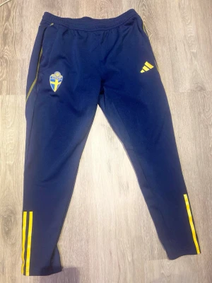 Blå Sverige Adidas träningsbyxor - Mörkblå träningsbyxor från Adidas med Sveriges landslagsemblem på ena benet och gula Adidas-detaljer. Byxorna har smal passform, resår i midjan och gula ränder längs benen. Perfekta för fotbollsträning eller gymmet.