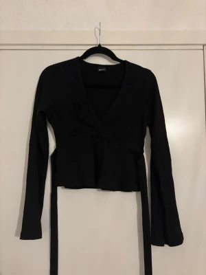 Svart knyt topp med lång ärm - Svart omlottopp från Gina tricot med långa ärmar och knytband i midjan. Toppen har en v-ringad design och är croppad, vilket ger en snygg och modern look. Perfekt att matcha med jeans eller kjol för en cool stil.