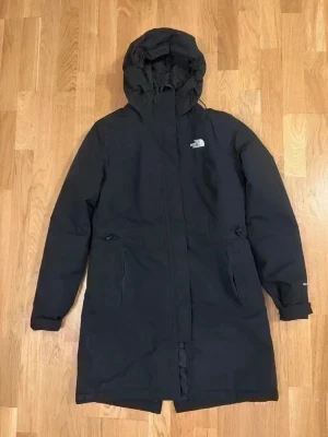  The North Face zaneck parka - Svart parkas från The North Face med huva och dragkedja framtill. Jackan har två stora fickor med lock och diskret logga på bröstet och ryggen. Lång modell som håller dig varm under kalla dagar. Perfekt för dig som vill ha en stilren och funktionell vinterjacka.