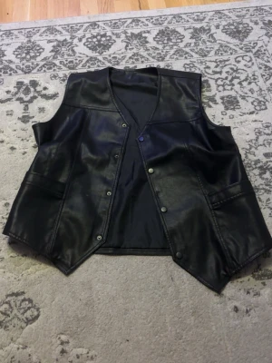 Svart läderväst - Säljer en svart läderväst med tryckknappar framtill och två fickor på sidorna. Västen har en rak passform och V-ringning, perfekt för att addera en cool touch till din outfit.