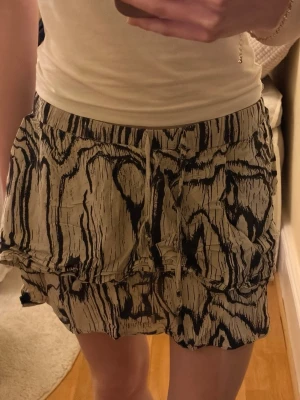 Beige och svart volangkjol med snörning - Kort kjol i beige med svart abstrakt mönster och volangkant. Kjolen har snörning i midjan och är gjord i ett lätt, luftigt material som känns skönt på sommaren. Passformen är normal och modellen är avslappnad. Storleken är M men liten i storlek, närmare S.