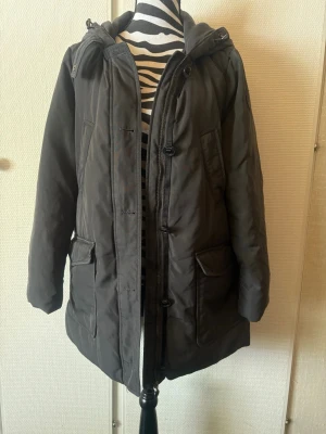 Svart parkas med huva - Säljer en svart parkas med huva och dragkedja samt knappar framtill. Jackan har två stora fickor med lock och två snedställda bröstfickor. Lång modell som passar perfekt när det är kallt ute. Fodrad för extra värme och har en stilren look.
