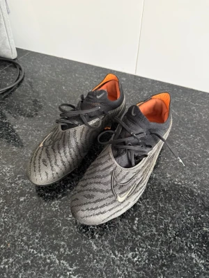 Nike Phantom GX Elite fotbollsskor FG - Snygga Nike Phantom GX Elite fotbollsskor i grått och svart med mönstrad ovandel och orange insida. Skorna är använda max 10 gånger och säljs då jag har lagt ner med fotbollen.