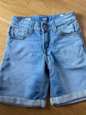 Ljusblå jeansshorts från Jack & Jones 8år - Säljer ett par ljusblå jeansshorts från Jack & Jones med klassisk femficksdesign, uppvikta benslut och bälteshällor. Shortsen har knappgylf och en snygg läderpatch med logga bak i midjan. Perfekta för varma dagar! Storlek 8år -128