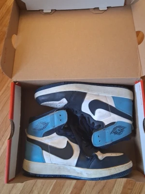 Nike Air Jordan 1 blå och vita sneakers - Säljer ett par Nike Air Jordan 1 sneakers i blått, vitt och svart. Skorna har högt skaft, klassisk swoosh-logga på sidan och Air Jordan-logga vid ankeln. Ovandelen är i läder och sulan är blå med bra grepp. Perfekta för dig som gillar streetwear och ikoniska modeller.