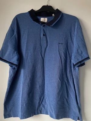 Blå pikétröja från GANT 4xl  - Klassisk blå pikétröja från GANT med mörkblå krage och ljusblå detaljer. Tröjan har korta ärmar, knappar framtill och diskret GANT-logga broderad på bröstet. Perfekt till jeans eller shorts för en avslappnad look. Storlek 4XL