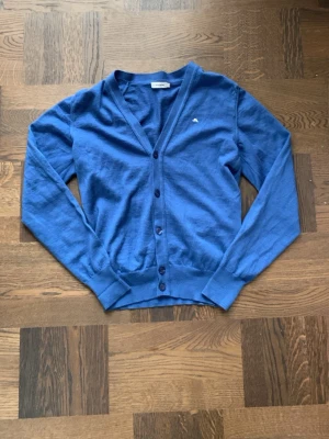 Mörkblå kofta med knappar från J.lindeberg  - Säljer en riktigt snygg stilren blå kofta från J.Lindeberg med knäppning framtill och långa ärmar. Klassisk passform och ribbade muddar vid ärmslut och nederkant. Passar lite åt S hållet. Vid funderingar, fråga på! 