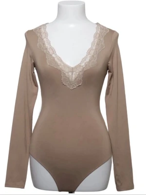 Beige långärmad body med spets - Snygg beige body med långa ärmar och djup v-ringning dekorerad med spetsdetaljer. Tight passform som framhäver siluetten och ger en feminin look. Perfekt att styla med jeans eller kjol för en trendig outfit.