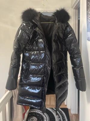 Glansig svart lång dunjacka med pälshuva - Snygg svart lång dunjacka med glansig finish och stor huva med svart fuskpäls. Jackan har quiltad design, dragkedja och tryckknappar framtill samt två fickor. Perfekt för kalla vinterdagar och ger en trendig look.