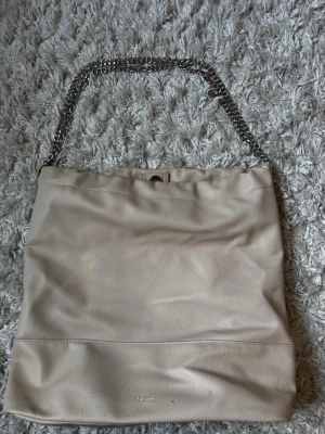 Rizzo väska - Snygg beige handväska i mjukt konstläder med silverfärgad kedjerem. Väskan har en enkel, stilren design och stängs med magnetknapp. Till väskan hör en matchande liten clutch i samma material. Perfekt för dig som gillar minimalistisk stil.