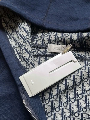 Marinblå hoodie från D1or - Exklusiv marinblå hoodie från D1or med ikoniskt D1or-mönster på insidan och broderad logga på fickan. Hoodien har dragkedja, huva och känguruficka. Tillverkad i mjukt material som känns lyxigt och bekvämt. Perfekt för dig som vill ha något stilrent och trendigt.