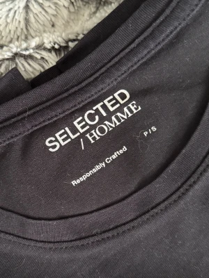 Svart t-shirt från Selected Homme - En stilren svart t-shirt från Selected Homme med klassisk rund hals. Tillverkad i mjuk bomull för skön känsla och enkel att matcha med allt. Perfekt basplagg för dig som gillar minimalistisk och clean stil.